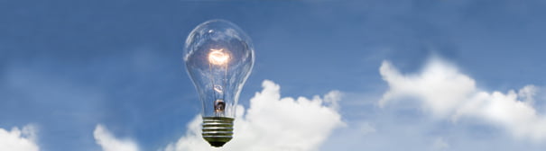 A lit lightbulb floating in front of a blue sky with a large puffy white cloud.